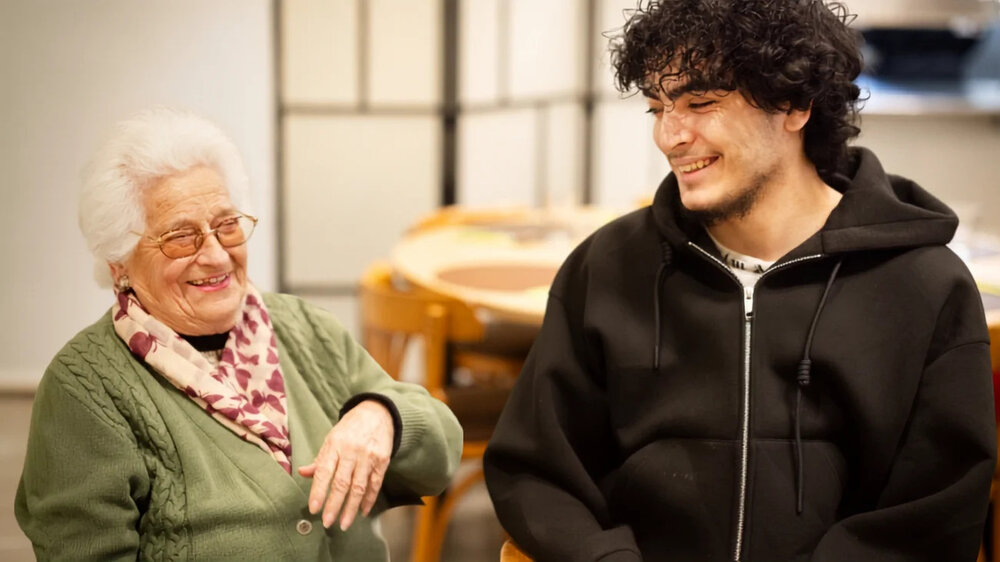 Jonge migrant samen aan het lachen met bejaarde vrouw.