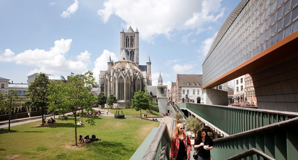 Zicht op de groene stadstuin rond de Sint-Niklaaskerk in Gent, met bezoekers die ontspannen in het gras, naast het moderne gebouw De Schaapstal onder een blauwe hemel met wolken.